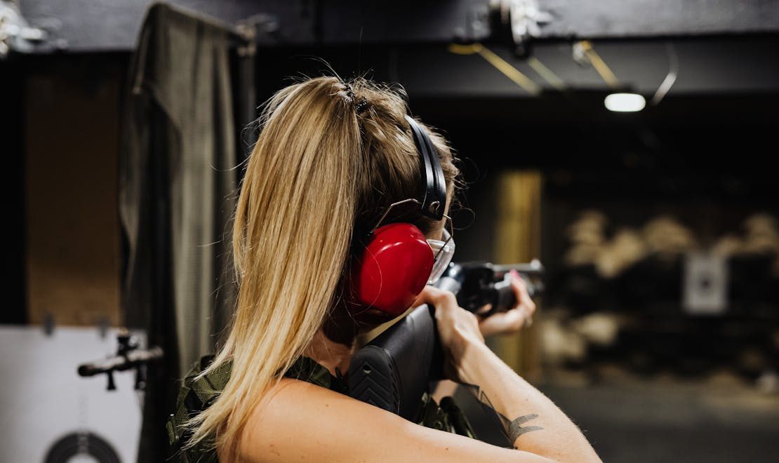 A person with long hair, wearing shooting earmuffs, aims a firearm in a shooting range, focused on a distant target.