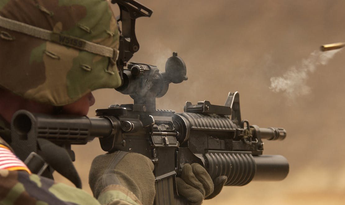 AR-15 vs AK-47 comparison showing a camouflaged soldier aiming an assault rifle as smoke rises from the barrel and a casing ejects.