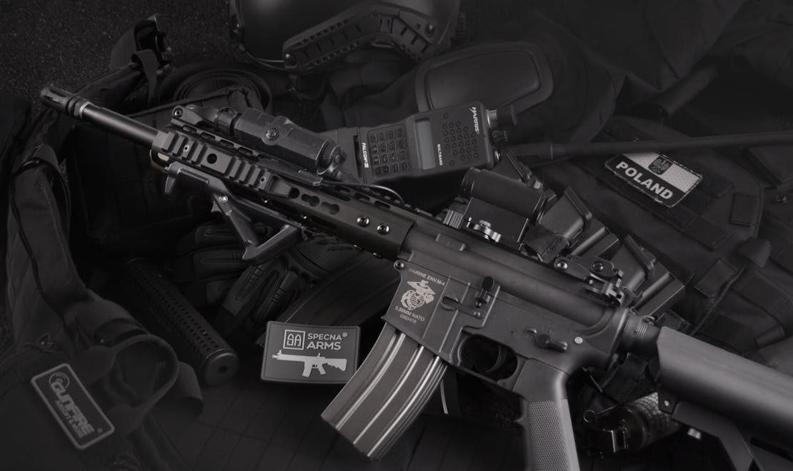 An assault rifle lying on tactical gear, including gloves, a radio, and a patch labeled "Poland," against a dark background.