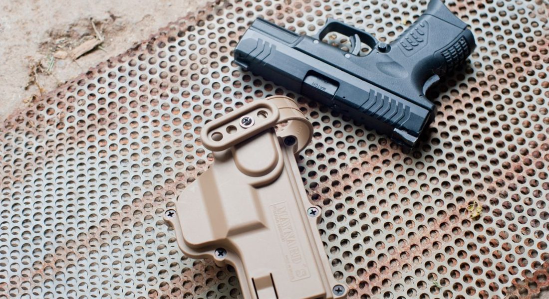 a top view of a black pistol beside a beige holster on a perforated metal surface outdoors