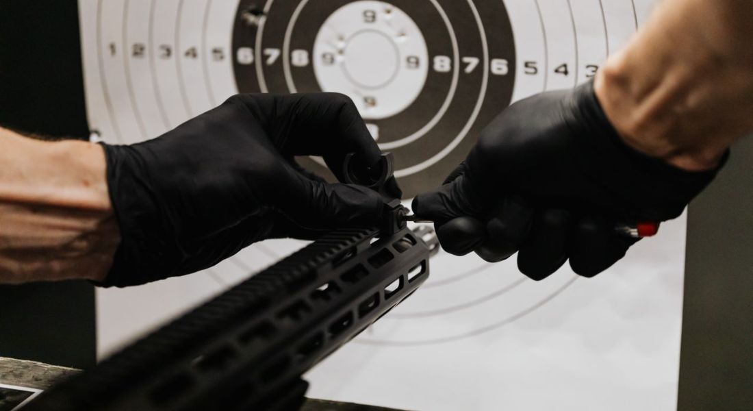 gloved hands adjusting a rifle's iron sight with a tool at an indoor shooting range