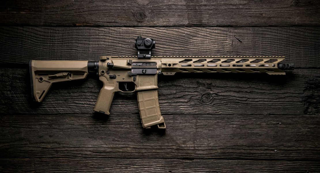 Tan AR-15 rifle with a red dot sight lying on a dark wooden surface