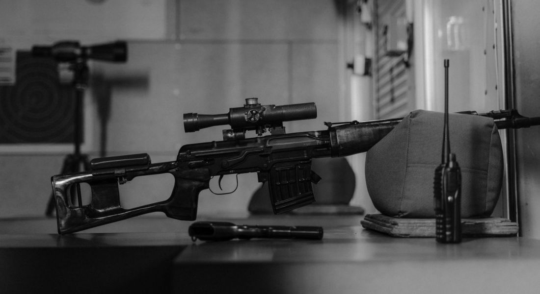 a monochrome image of a sniper rifle on a table indoors with a walkie talkie and scope